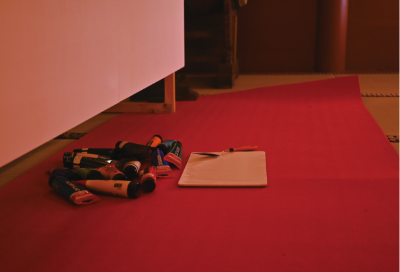 大きな白いキャンバスの前、赤い絨毯に置かれたアクリル画材と白いパレット